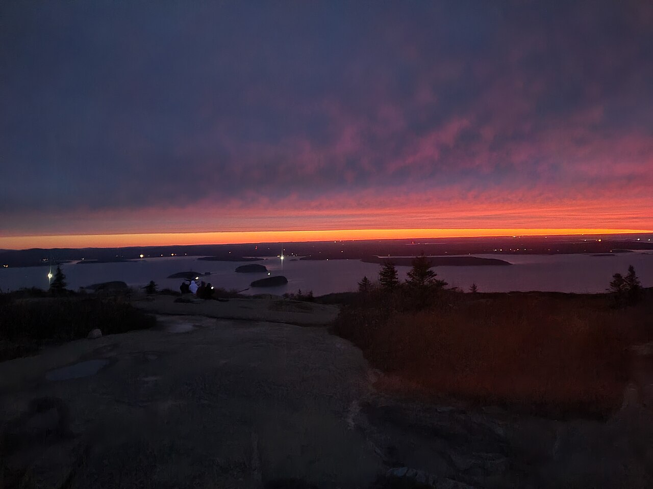 The sun rising over the Atlantic Ocean and coastal islands, casting golden light across the granite summit of Cadillac Mountain in Acadia
